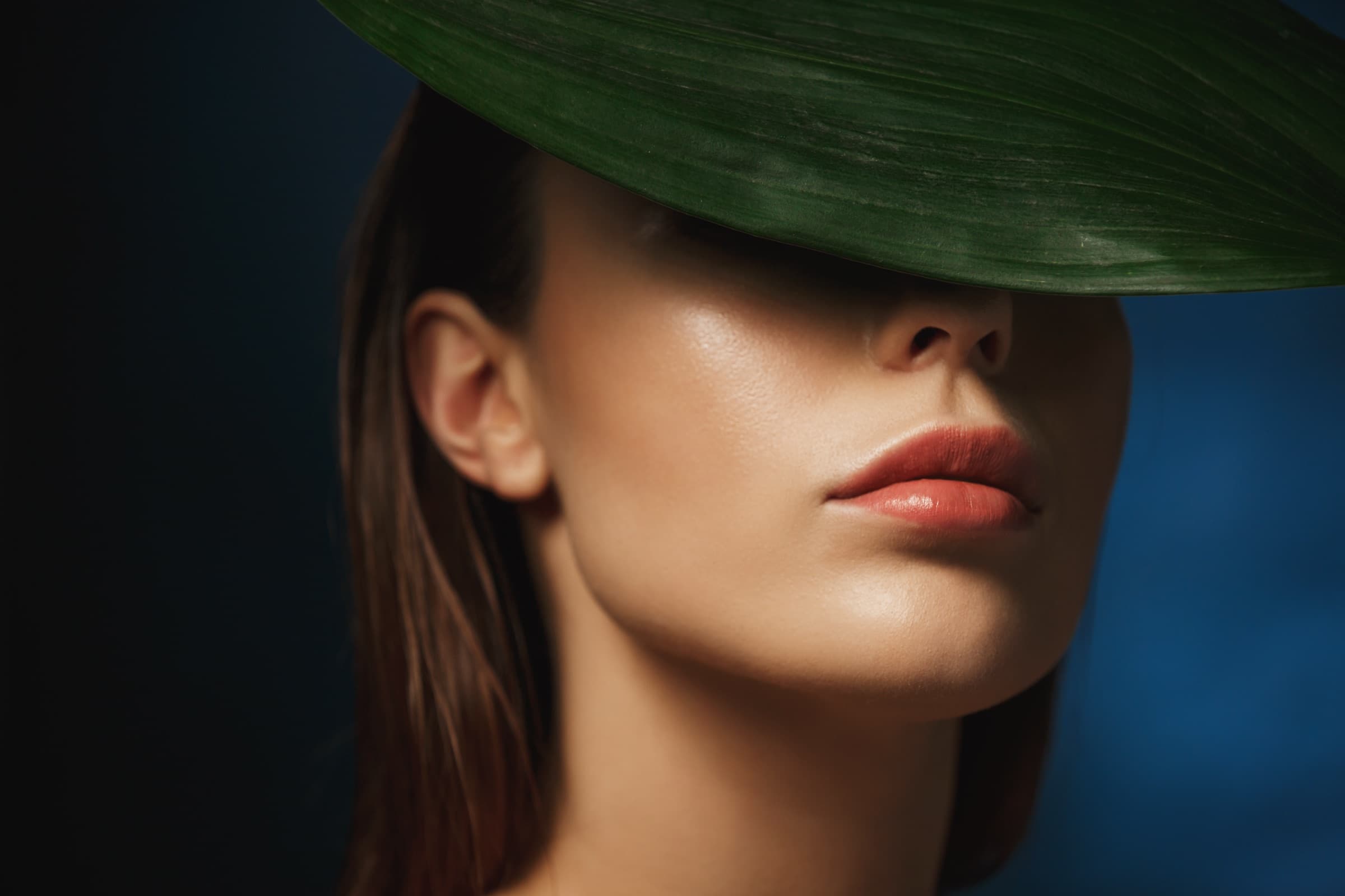 A model's lower face framed by a single tropical leaf against a deep blue backdrop — a Bespoke Beauty editorial portrait.