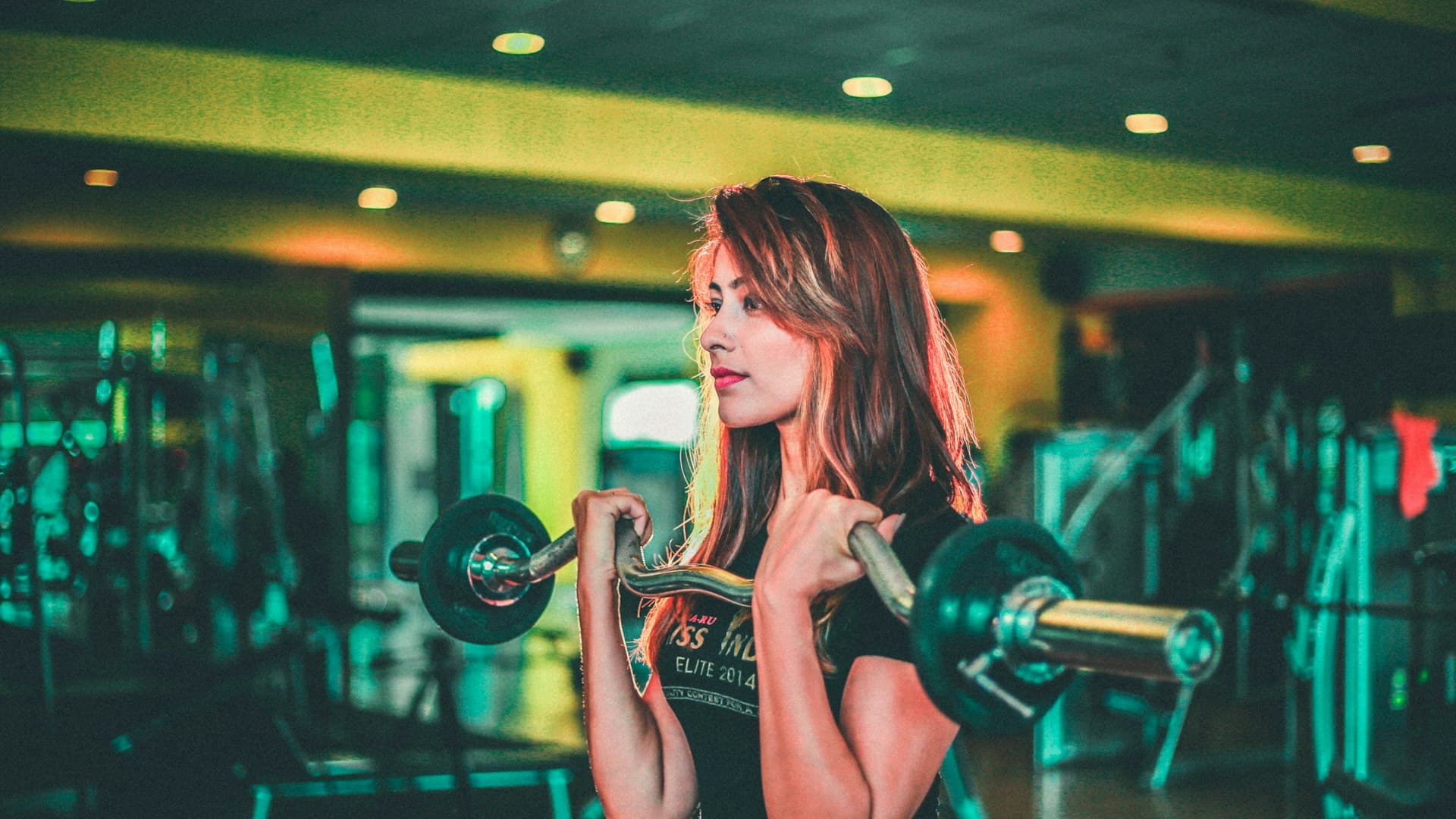 Seorang perempuan mengangkat barbel hitam dan abu-abu dengan postur kuat di gym