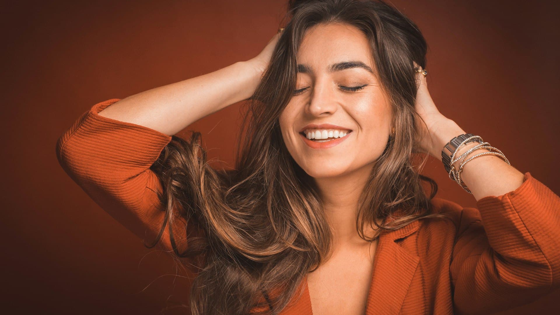 Perempuan tersenyum mengenakan atasan cokelat sambil memegang helai rambutnya, memeriksa kepadatannya