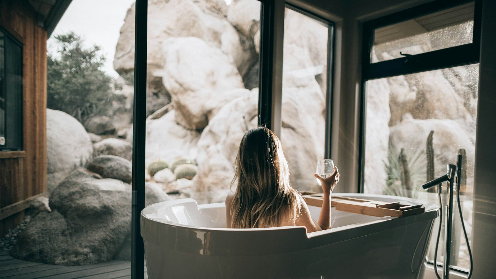 Perempuan berendam santai di bathtub putih sambil memegang gelas air bening