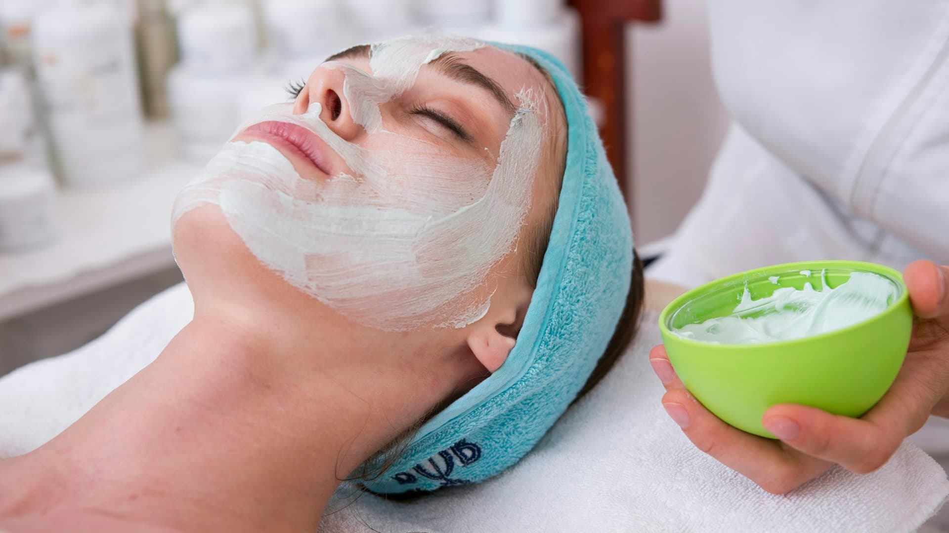 Perempuan berbaring di handuk biru dengan krim putih di wajah saat perawatan
