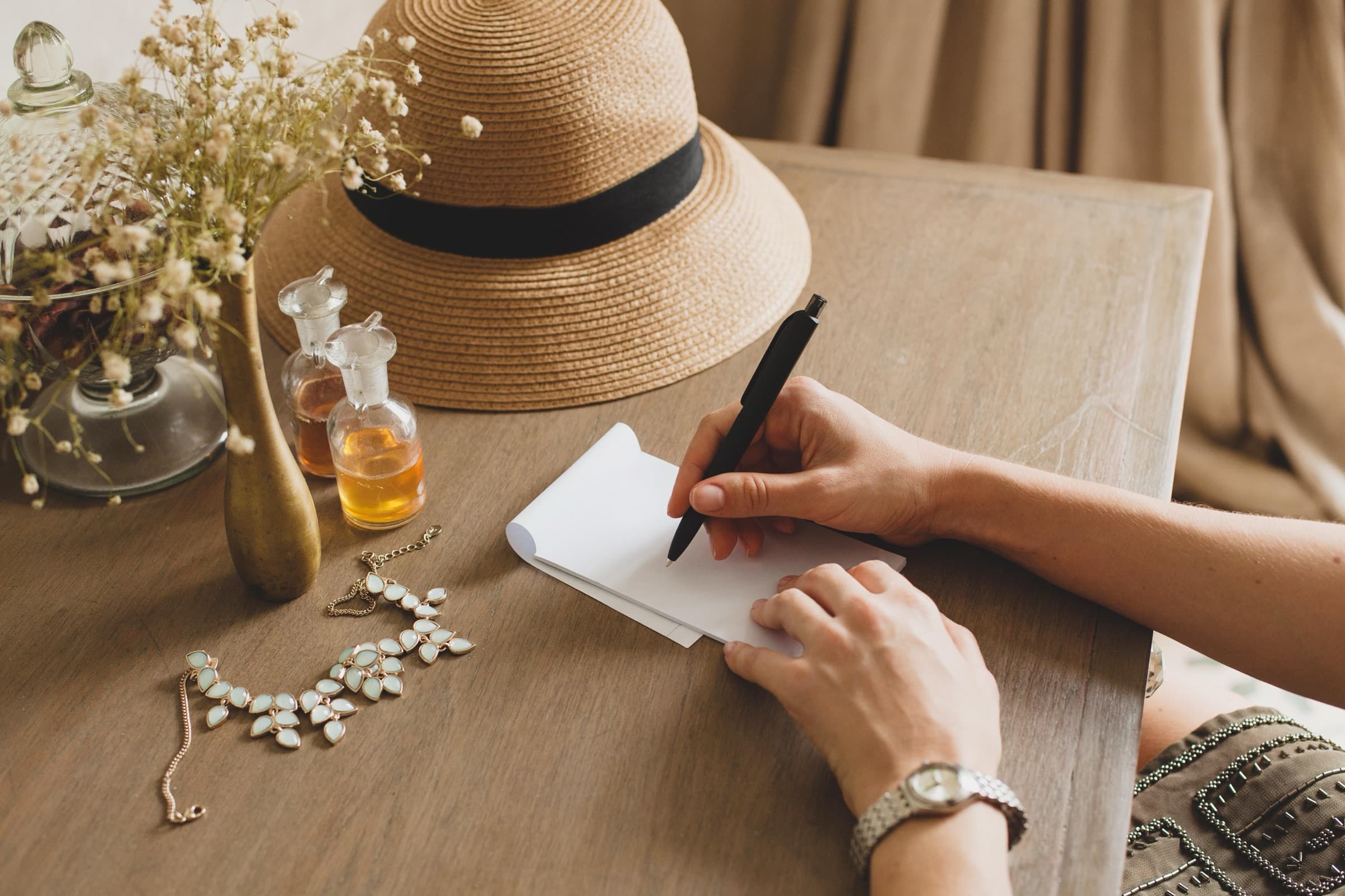 A writer composing notes at a styled desk with a straw hat, perfume, and jewelry — a Bespoke Beauty editorial scene.