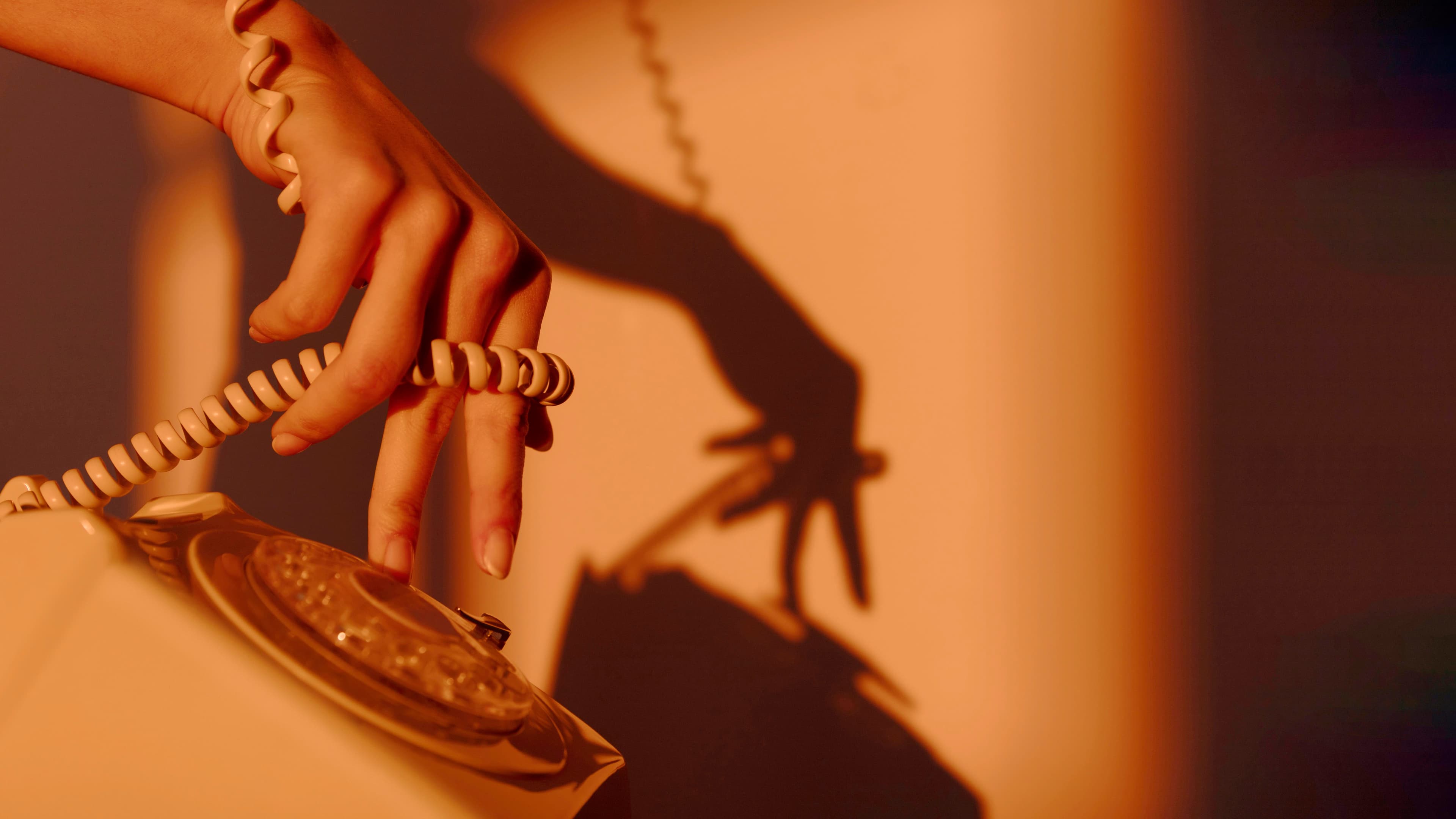 A hand lifting the receiver of a vintage rotary telephone, casting a dramatic silhouette against a warm amber wall — a Bespoke Beauty editorial contact scene.