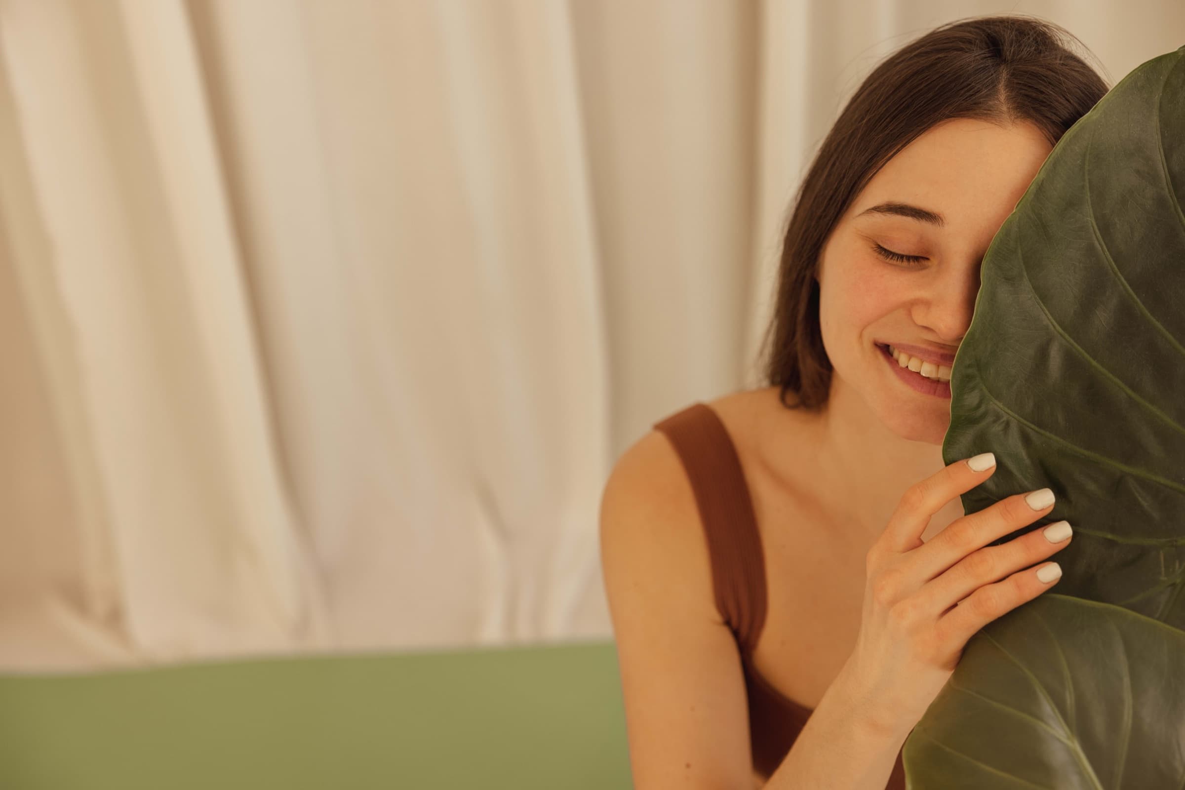 A smiling woman resting her cheek against a large green leaf in soft natural light — a Bespoke Beauty editorial portrait.